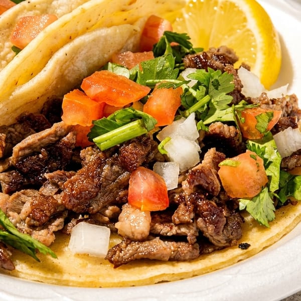 Two corn tortilla tacos with grilled meat, diced tomatoes, onions, cilantro, and a lemon wedge.